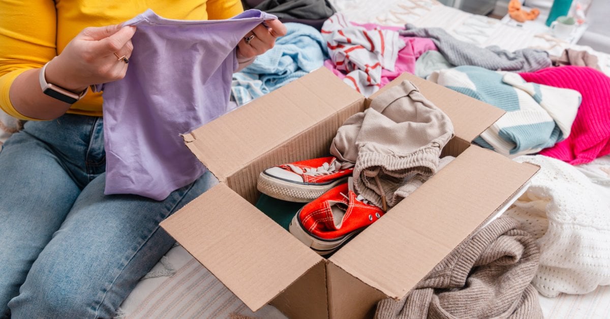 A person sits on a bed and holds a purple top before her. An open cardboard box on the bed has clothes and red shoes inside.