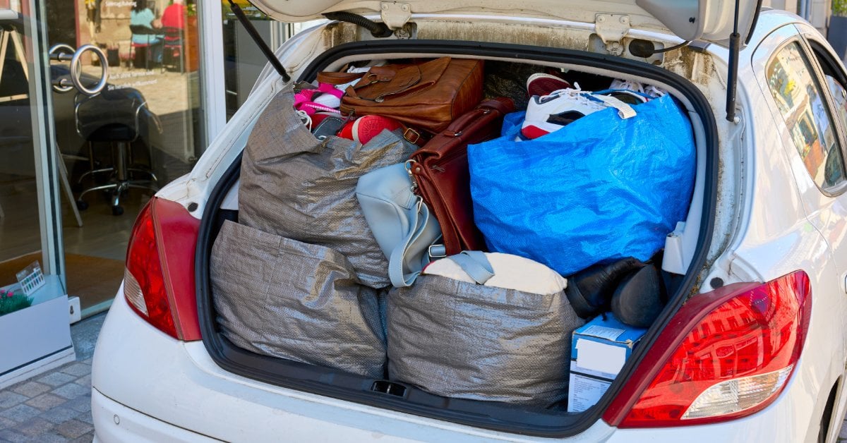 The trunk of a white SUV is open and filled with three gray toes, a large blue bag, and brown purses.