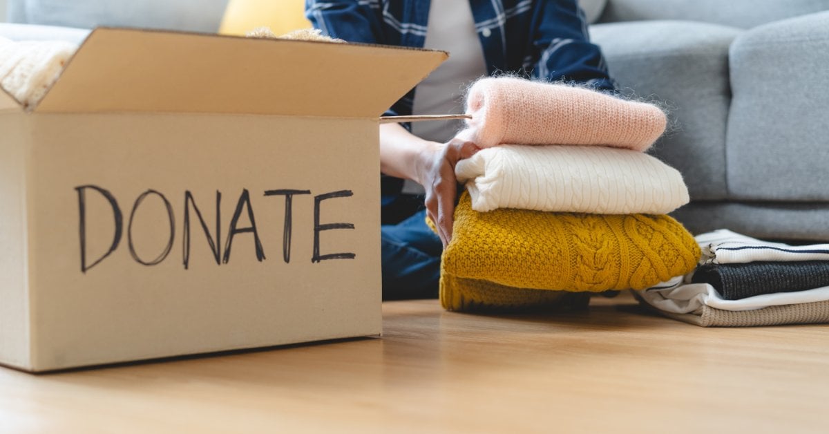 A woman sits on the floor in front of a couch. She's folding sweaters beside an open cardboard box labeled "DONATE."